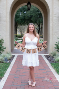 A senior girl in a white dress is holding a 'senior' banner while Khim Higgins Photography takes her photo.