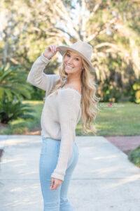 A beautiful blond high school senior wearing a hat and a sweater  turns and smiles over her shoulder looking at the camera. 