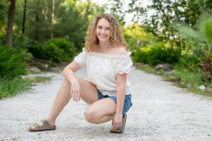 A senior girl wearing a white top and blue jean shorts is squatting down in a pose for Khim Higgins Photography.