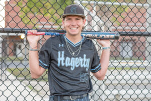 senior boy in a Hagerty High School baseball uniform is holding a bat and leaning against a fence