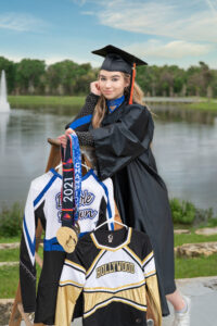 A senior girl wearing a cap and gown is standing on a ladder holding her cheer medals.