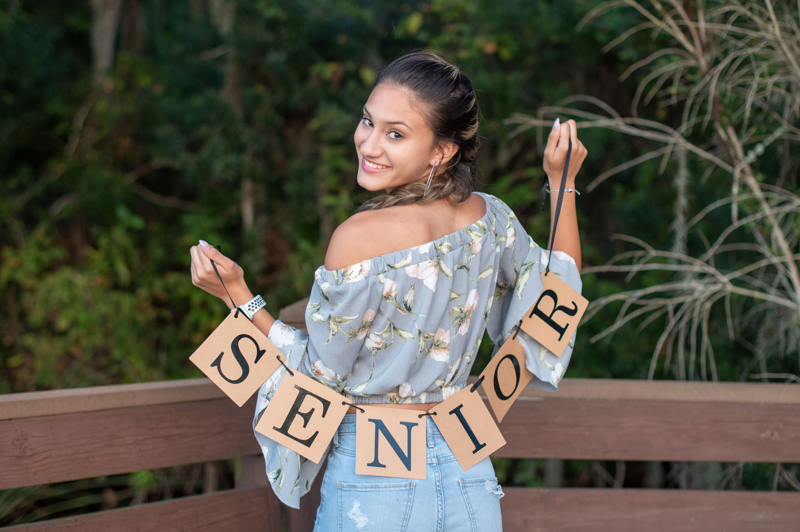 A beautiful high school senior girl holding a 'senior' banner sign behind her back and looking back at the camera smiling.