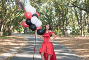 A girl wearing a red formal dress is holding balloons and smiling at Khim Higgins Photography for her sweet 16 photo shoot.