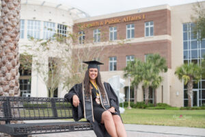 A UCF graduate girl is sitting on a bench in front of the business administration building while Khim Higgins Photography takes her photos.