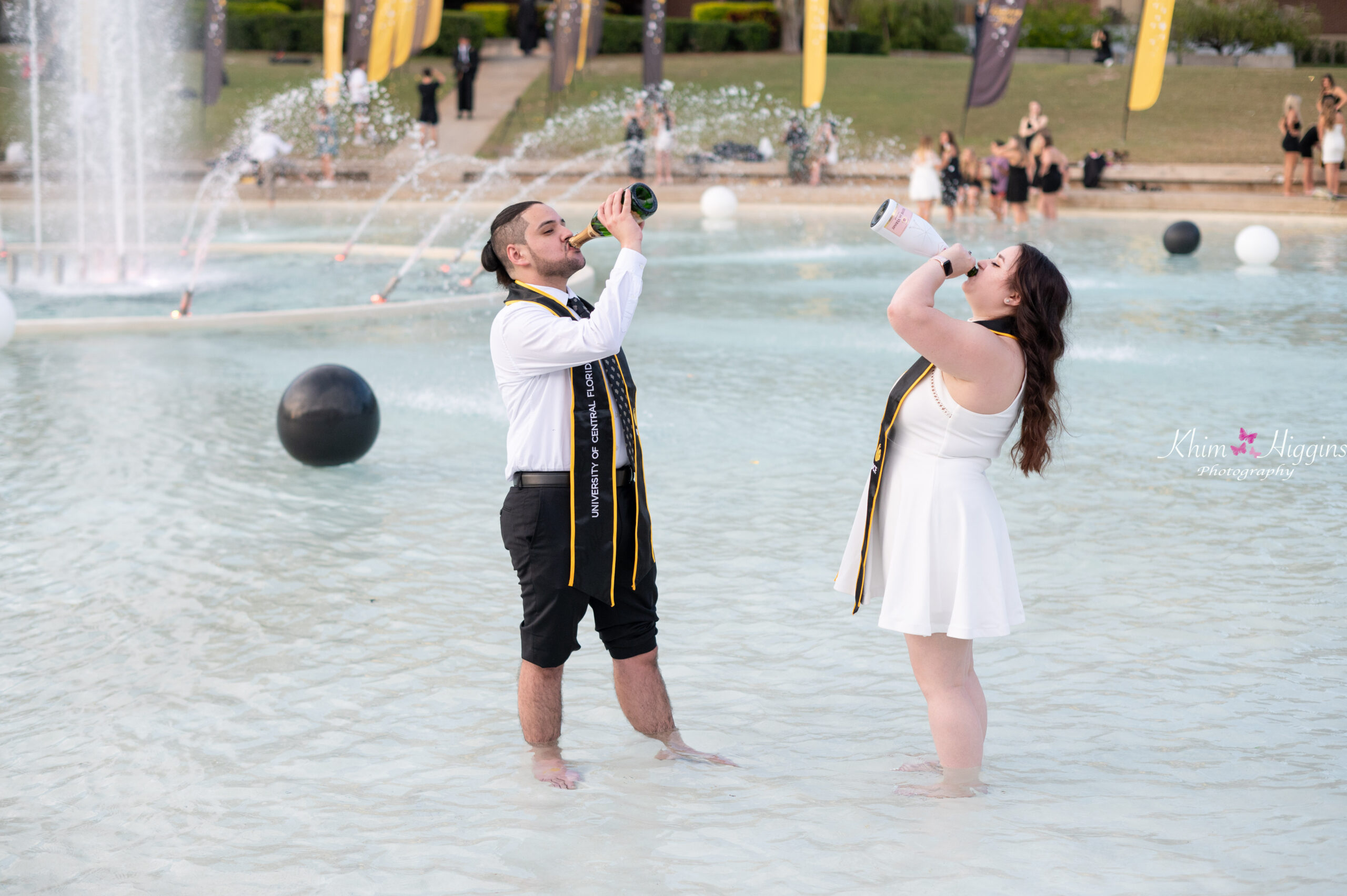 A guy and a girl standing in a fountain drinking a bottle of champagne while UCF Graduation Photographer Khim Higgins takes their photos.