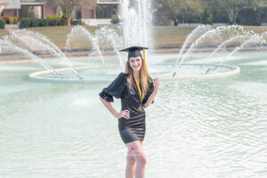A UCF graduate girl is wearing a black dress with a graduation cap is standing in the fountain while Orlando UCF graduation photographer takes her photos.