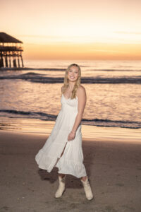 A high school senior girl in a white dress with boots on is swaying back and forth on the beach smiling at Orlando senior photographer, Khim Higgins during a sunrise session.