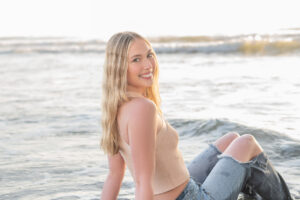 A high school senior girl is wearing jeans and a tank top is sitting in the water at the beach and looking over her right shoulder at Orlando Senior Photographer, Khim Higgins during a sunrise session at Cocoa Beach, Florida.