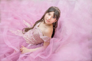 A girl wearing a pink sweet 16 formal gown is sitting on stairs and looking up at Khim Higgins Photography.