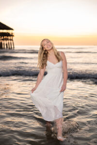 A high school senior girl wearing a white dress is walking on the beach and smiling at Khim Higgins Photography.