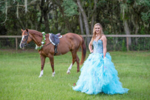 A sweet 16 girl is wearing a blue formal dress with a horse behind her while Khim Higgins Photography photographers her sweet 16 picture.