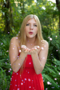 A blond girl in a sleeve less dress is blowing flower confetti while Khim Higgins Orlando Senior Photographer takes her picture.