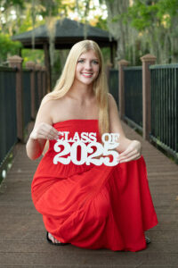 A senior girl wearing a red dress is holding a class of 2025 sign while Khim Higgins Photography takes her picture.