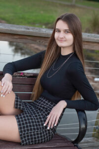 Lake mary senior photo shoot with a senior girl sitting on a bench by the lake. She is wearing a long sleeve black shirt and a short black checkered skirt.