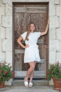 Lake Mary high school senior girl wearing a short white dress is posing in front of a brown door with one arm on the door and one hand on her waist.