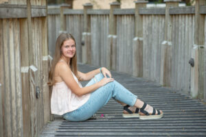 Jen, a senior at Lake Mary High School is wearing jeans and a pink tank top sitting at the dock leaning on the side of the wood.