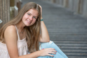 Lake mary senior girl in a pink tank top with blue jeans is sitting at a dock with her left hand touching her hair.