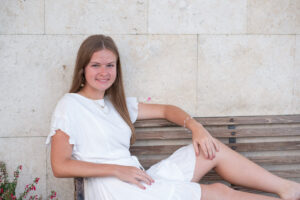 lake mary senior girl in a short white dress is sitting on a bench and leaning on one side of the bench arm smiling at Khim Higgins Photography.