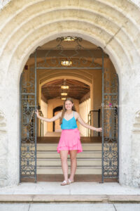 Jen, a lake mary senior is wearing a blue tank top and pink skirt is standing in the center of a rot iron archway with her arms stretched out touching the archway.