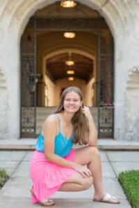 Lake mary senior girl wearing a blue tank top and pink skirt is squatting down in front of a beautiful arch smiling at Khim Higgins Photography.