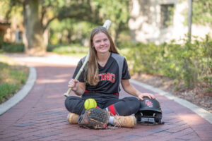 Lake Mary Senior Girl photo shoot with the senior girl sitting on the sidewalk holding a baseball bat over her right shoulder and a baseball glove, ball and helpmet on the floor next to her legs.