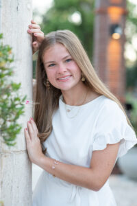 Lake mary senior girl photo shoot with the model wearing a short white dress and she is leaning against a white pillar with a beautiful smile.