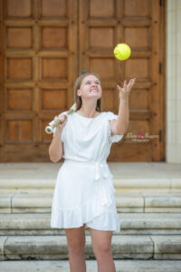 Lake mary senior girl photo shoot with a senior wearing a white dress holding a baseball bat over her right shoulder and she is tossing a softball up in the air.