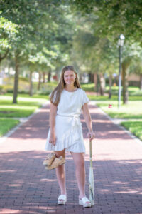 Lake mary senior girl photo shoot with a senior girl in a white short dress holding her baseball shoes in one hand and leaning on a baseball bat with the other hand.