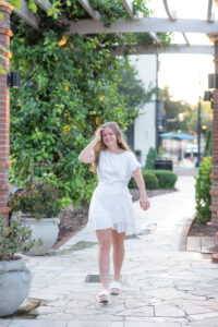 Lake Mary senior girl in a short white dress is walking on a side walk with her right hand sweeping her hair back.