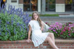 Lake Mary Senior Photo shoot with Jen who is wearing a short white dress sitting around a bunch of purple and pink flowers.