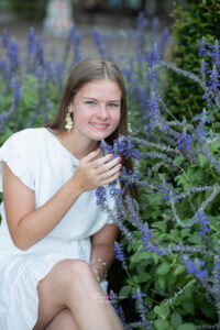 lake mary senior girl in a white dress is sitting near a bunch of purple flowers.