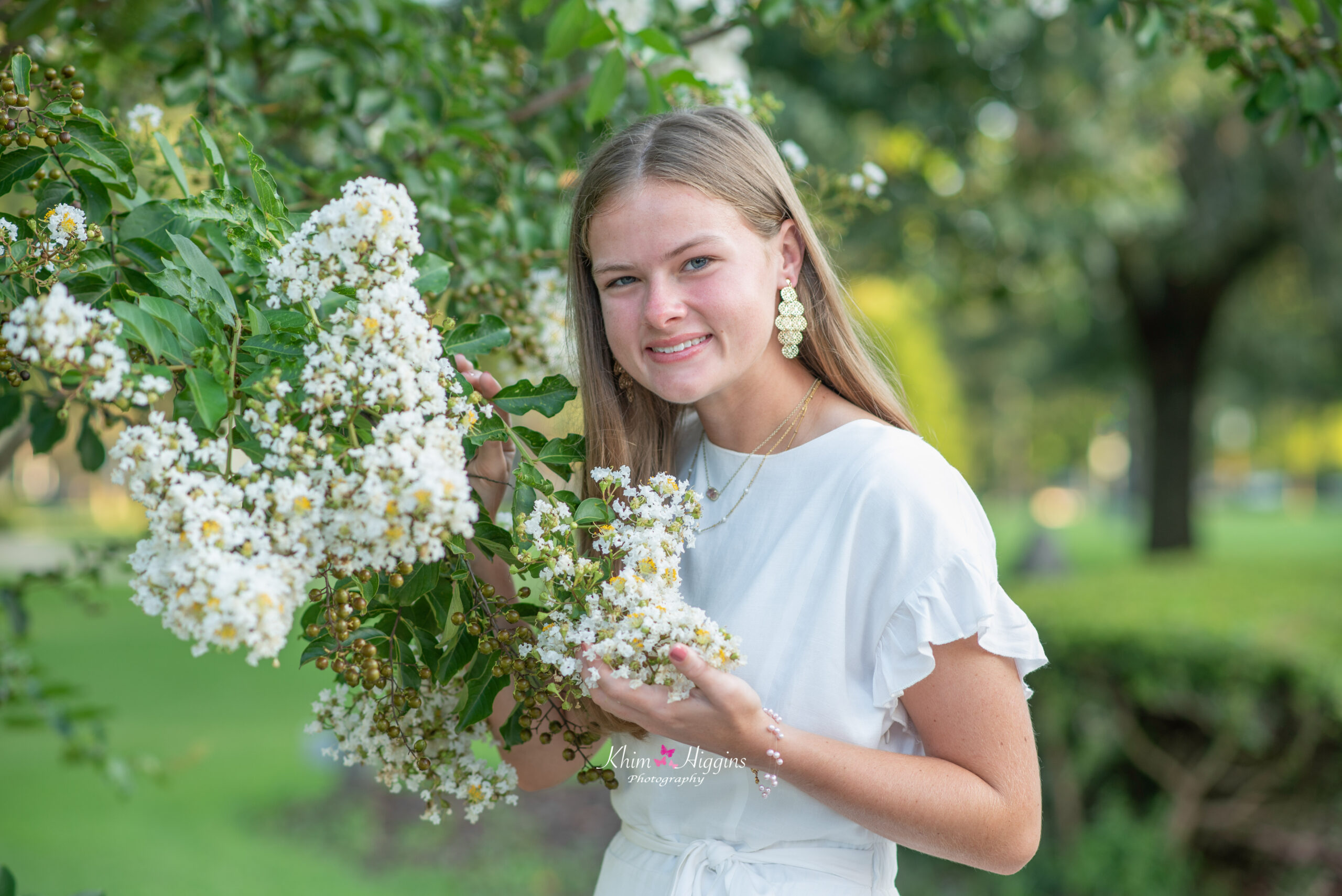 Lake mary senior photo shoot with a senior girl wearing a white dress and she is standing by a tree and holding white flowers that are hanging from that tree.