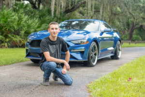 An Oviedo High School Senior is wearing a black t-shirt and jeans is kneeling in front of his blue sports car while Khim Higgins Photography takes his photo.