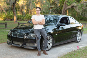 A high school senior boy wearing a white t-shirt and black jeans is leaning against a black BMW car smiling at Khim Higgins Photography, Orlando Senior Photographer.