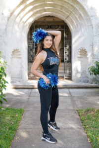 A senior girl wearing her school dance uniform and holding blue pom poms in each hand.