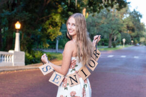 senior girl holding a banner 'senior' over her shoulders and turning around and smiling at the photographer, Khim Higgins.