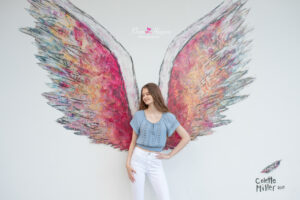 Lake Mary senior photo shoot with Ella, a senior who is wearing a blue top and white jeans is standing in front of a mural painting of angel wings.