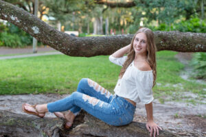 Oviedo senior girl in a white top with blue jeans is sitting on a branch at sunset at a park in downtown Orlando as Khim Higgins Photography takes her photos.