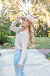 A senior girl in a cream color sweater and light jeans is wearing a hat and looking back at Khim Higgins Photography as she tips her hat and looks back over her left shoulder.