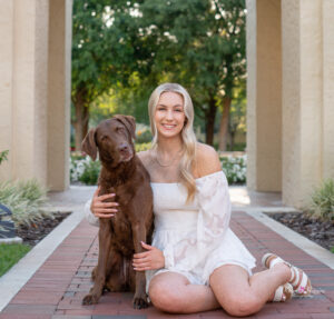 Hagerty high school senior girl is wearing a white dress and is sitting next to her chocolate lab.