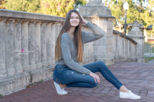 Lake Mary High School Senior Photo Shoot with a senior girl wearing jeans and a gray long sleeve shirt is squatting down on a bridge.