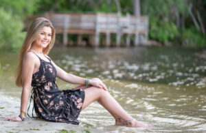 A senior girl wearing a black dress is sitting on sand by the lake.