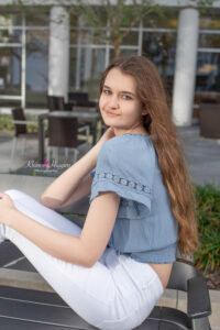 Lake mary senior photo session with Ella. Ella is wearing a blue short sleeve shirt with white jeans and she is sitting on a bench and looking over her left shoulder at Orlando senior photographer.