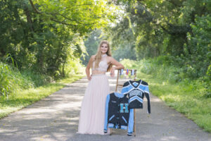 A senior girl from Hagerty High School is wearing a formal pink top and pink skirt leaning on a ladder that has her cheer uniform shirt and jacket hanging on the ladder.