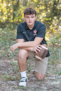 A senior boy is wearing a black shirt with khaki shorts is kneeling with one knee and holding a football.