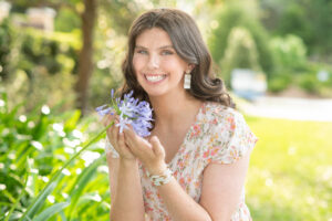 A high school senior girl holding a purple flower is smiling at Khim Higgins Photography.