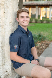 A senior boy in a dark blue shirt and khaki shorts is squatting down and leaning against a wall.