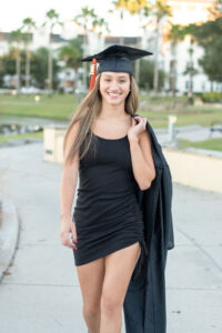 senior girl from Oviedo High School is wearing a short black dress and she has her grad cap on her head and holding her gown behind her shoulder.