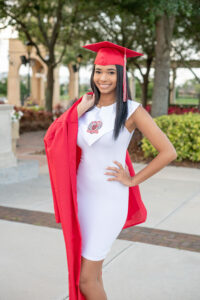 A senior girl in a white dress is wearing her high school graduation cap and holding her graduation gown over her right shoulder.