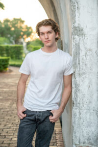 Senior boy is wearing a white t-shirt with dark jeans is leaning against a building